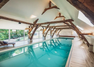 Piscine intérieure avec poutres en bois apparentes, entourée de grandes fenêtres offrant une vue sur la végétation extérieure.