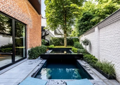 Chez-Anna-et-Luc-1 Vörös téglafalú ház mellett kis udvar medencével, fehér téglafal és zöld növényzet övezik, középen egy fa áll.