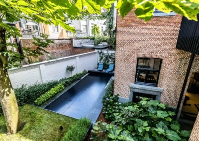 Chez-Anna-et-Luc-3 Téglaépület melletti kert, medencével és két kék napozóággyal, körülvéve zöld növényzettel és fehér kerítéssel.