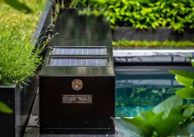 Chez-Anna-et-Luc-4 Fekete napelemes doboz egy medence mellett, körülötte zöld növények és cserjék.