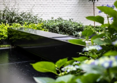 Chez-Anna-et-Luc-6 Fekete, fényes medencefedél zöld növényekkel körülvéve, fehér téglafal előtt.