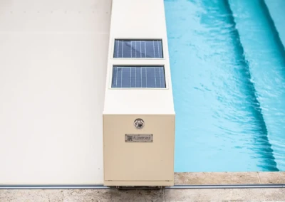 Fehér burkolatú medence takarórendszer napelemes panellel a medence mellett, kék víz és kőburkolat látható.