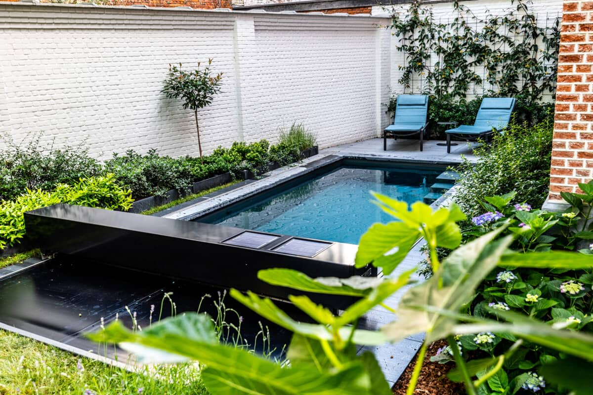 Két kék napozóágy egy kerti medence mellett, fehér téglafalak és zöld növények veszik körül.