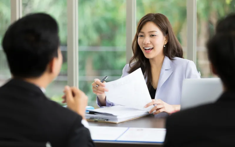 Két ember ül egy asztalnál, míg egy harmadik személy papírokat tart a kezében, háttérben zöld növények láthatók.