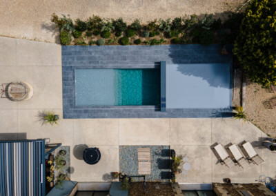 Egy téglalap alakú medence szürke burkolattal körülvéve, mellette napozóágyak és egy kerek asztal napernyővel.