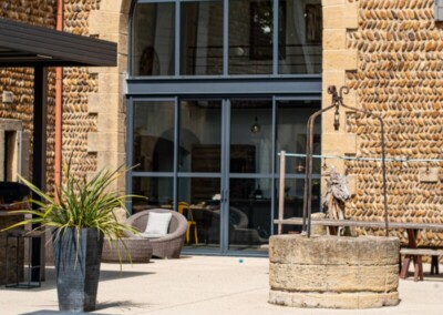 Régi kőépület íves ablakkal, előtte terasz, szürke medencefedéssel és növényekkel.