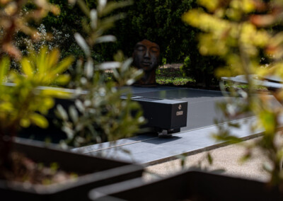 Kertben található szürke medencefedél körül növények, háttérben egy nagy szoborfej látható.