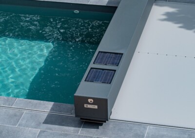 Szürke medencefedél napelemekkel, kék víz felett, körülötte szürke járólapok és zöld növények.