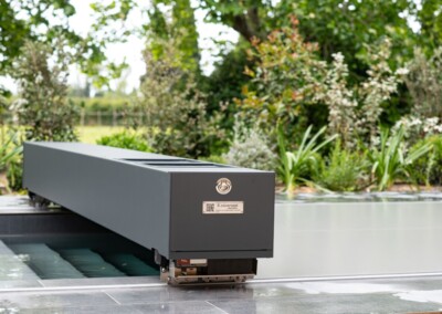 Szürke medencetakaró szerkezet zöld kert háttérrel, fémes részletekkel és növényekkel körülvéve.