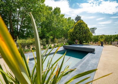 Kerti medence szürke burkolattal, körülötte zöld növények és fák. Az előtérben pálmalevelek láthatók.