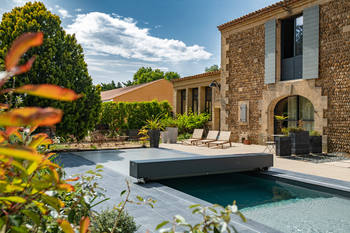 Kövezett terasz medencével és napozóágyakkal, háttérben kőfalú épület és zöld növényzet.