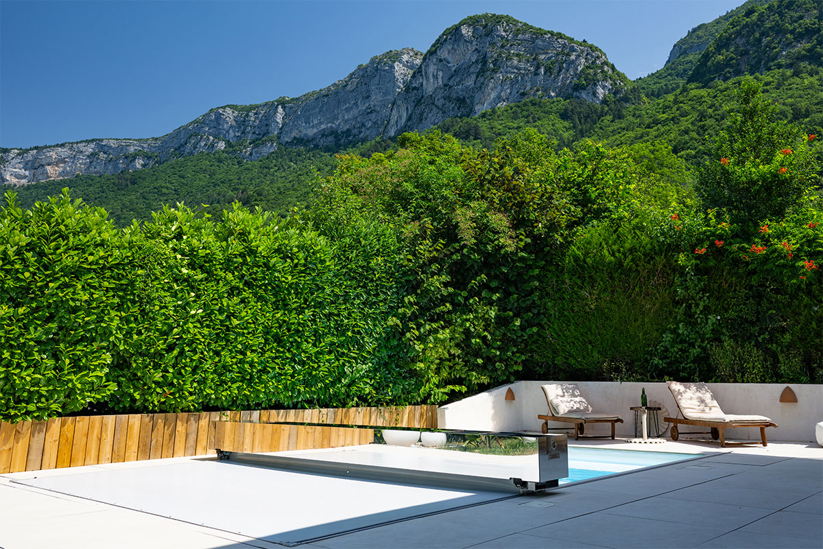 Medence fedéllel, napozóágyak és zöld növények előtt, háttérben hegyekkel és kék éggel.