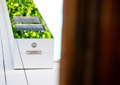 Coverseal-landscape5 Fém doboz tetején zöld növények tükröződnek, két napelemmel és egy záró mechanizmussal, fehér padlón.