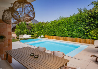 Coverseal-landscape7 Kerti medence mellett fa étkezőasztal székekkel, körülötte sövény és lámpák a teraszon.