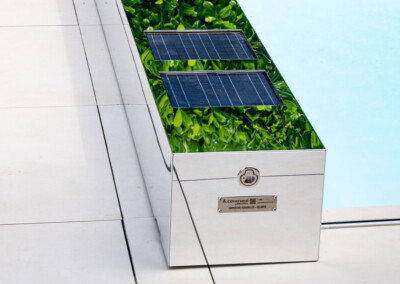 Coverseal-landscape8 A kép egy fényes fém dobozt ábrázol, amelyen napelemek és zöld levelek tükröződése látható, mellette világos csempe található.