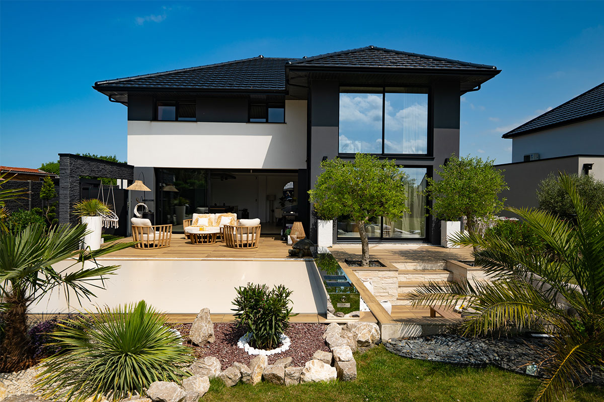 Kétszintes modern ház teraszán fa bútorok és zöld növények láthatók, kék égbolttal a háttérben.