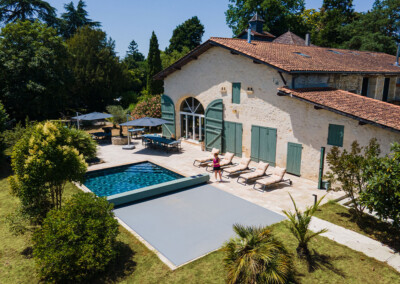 Egy vidéki ház udvarán medence, napozóágyak és zöld növények láthatók, körülötte fák és kerti bútorok.