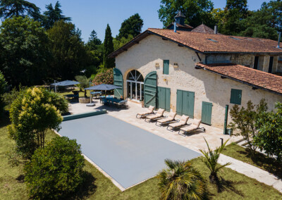 Kőház zöld zsalugáterekkel és napozóágyakkal szegélyezett terasszal, medencefedéllel, körülötte fák és bokrok.