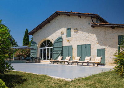 Kőház világos falakkal és zöld zsalugáterekkel, előtte napozóágyakkal és terasszal, körülötte zöld növényzet.