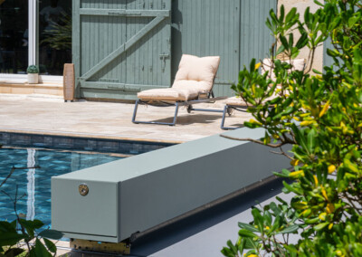 Napozóágyak krémszínű párnákkal egy medence mellett, zöld faajtó és növények a háttérben.