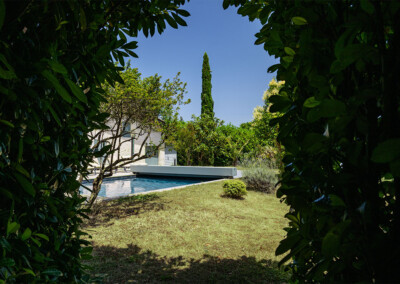 Kert medence mellett, buja zöld növényekkel körülvéve, háttérben egy ciprusfa és kék ég látható.