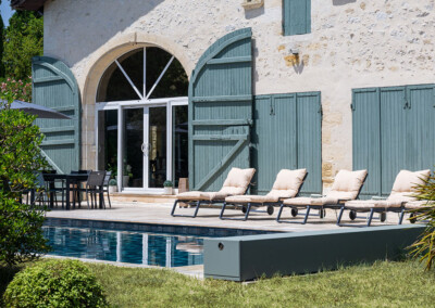Kerti medence mellett napozóágyak és asztal székekkel, háttérben kőház kék zsalugáterekkel.