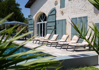 Négy bézs napozóágy egy medence mellett, zöld zsalugáteres épület előtt, napernyővel és asztallal a háttérben.