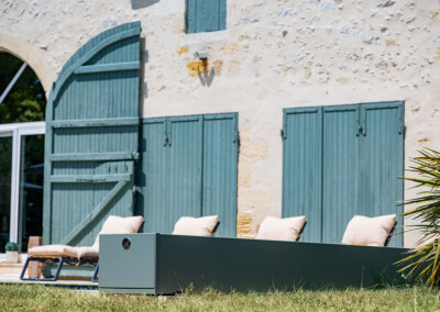 Kőház zöld zsalugáterekkel, előtte négy bézs párnával ellátott napozóágy és egy modern, szürke kültéri egység.