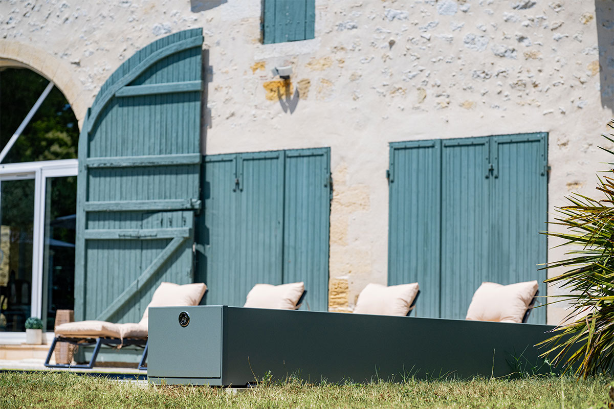 Kőház zöld zsalugáterekkel, előtte négy bézs párnával ellátott napozóágy és egy modern, szürke kültéri egység.