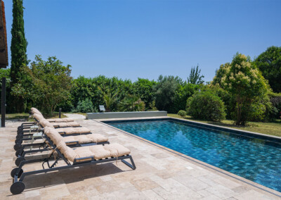 Kerti medence mellett hat napozóágy bézs párnákkal, körülvéve zöld növényzettel és kék ég alatt.